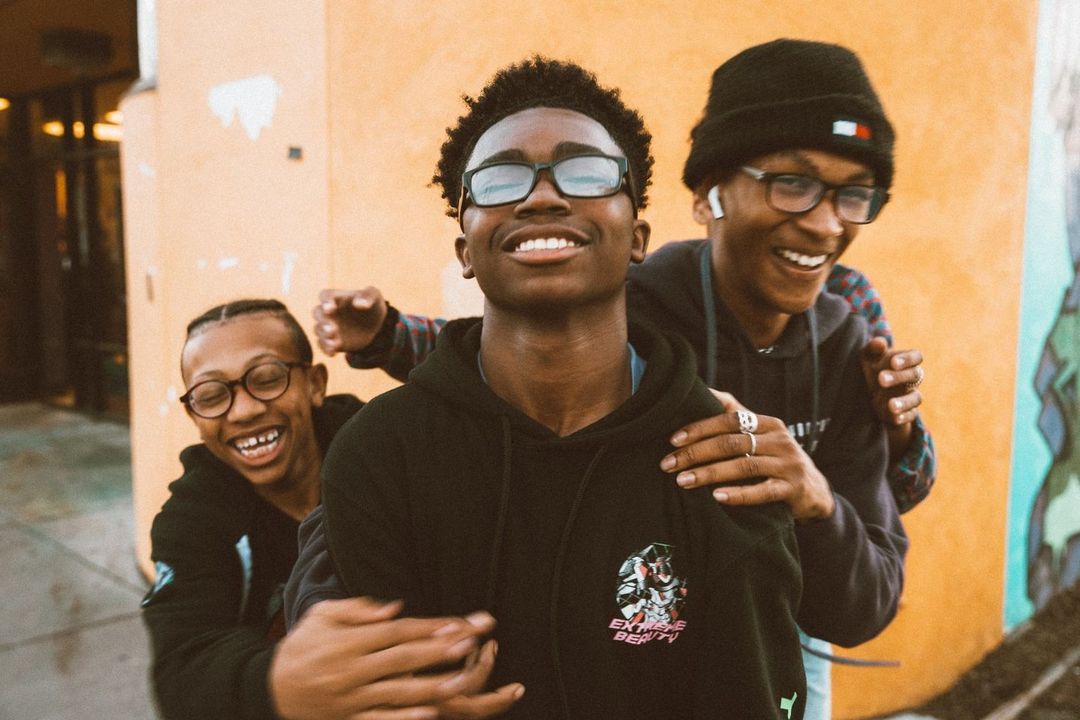 Three smiling African-American young adults pose outdoors against a yellow wall with graffiti; all wear glasses and casual clothing.