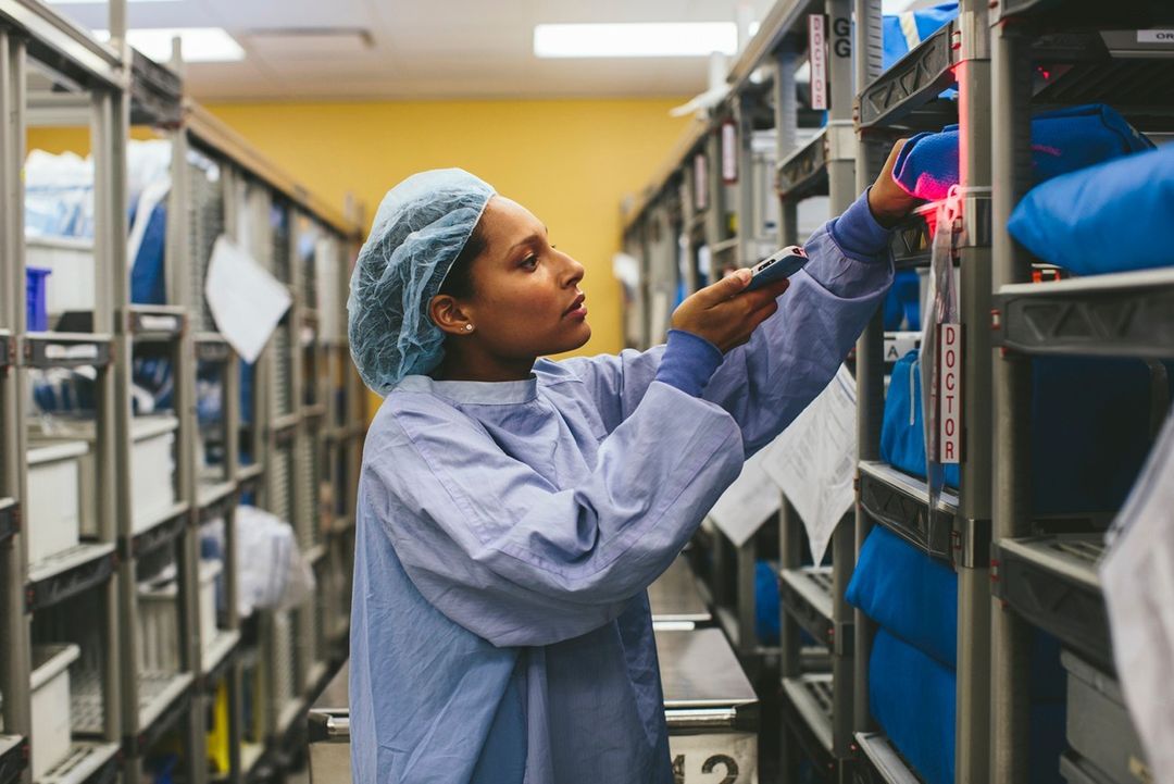 Healthcare worker scans inventory in a storage room. Shelves are stocked with blue wrapped surgical supplies. She wears scrubs and hair net.