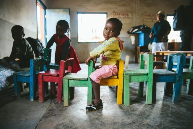 Young girl sits on a colorful chair in classroom surrounded by other children and chairs, creating a candid scene of early education.