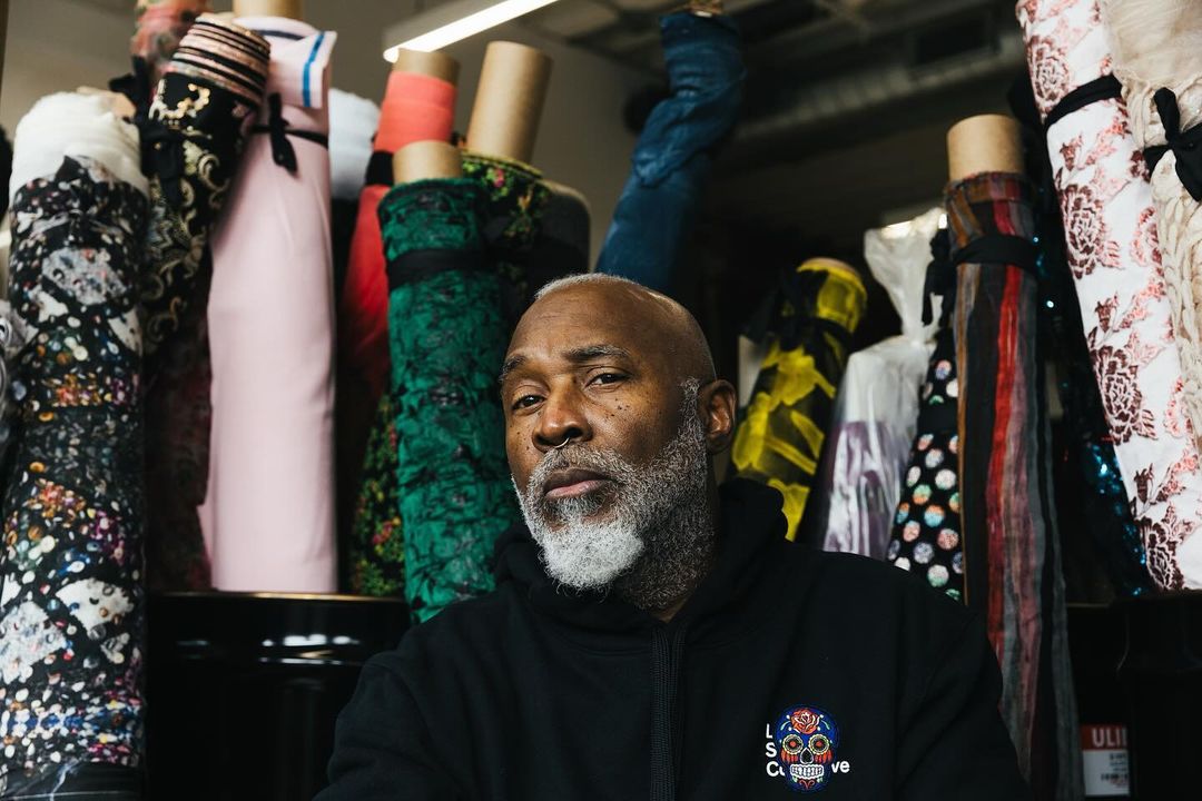 Adult male with grey beard in black hoodie poses indoors in front of bolts of colourful printed fabric, looking at the camera.