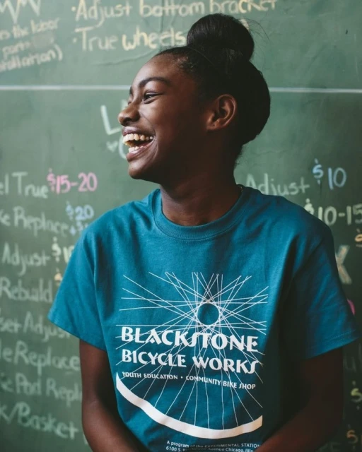 Teen smiles in a close-up photo wearing a Blackstone Bicycle Works t-shirt with handwritten chalkboard notes in the background.