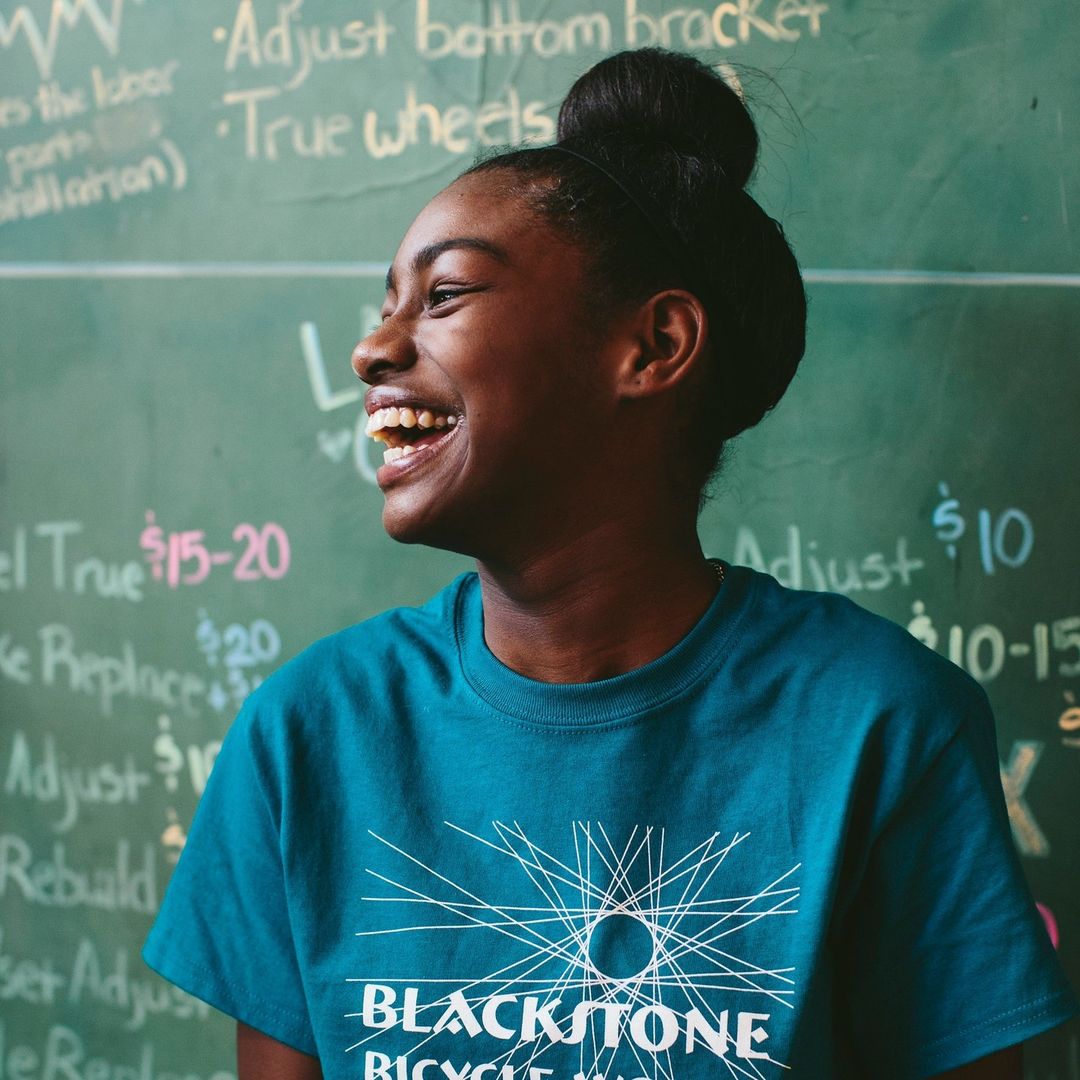 Smiling young Black woman wearing a turquoise t-shirt with a bicycle wheel logo against a green chalkboard with repair prices listed.