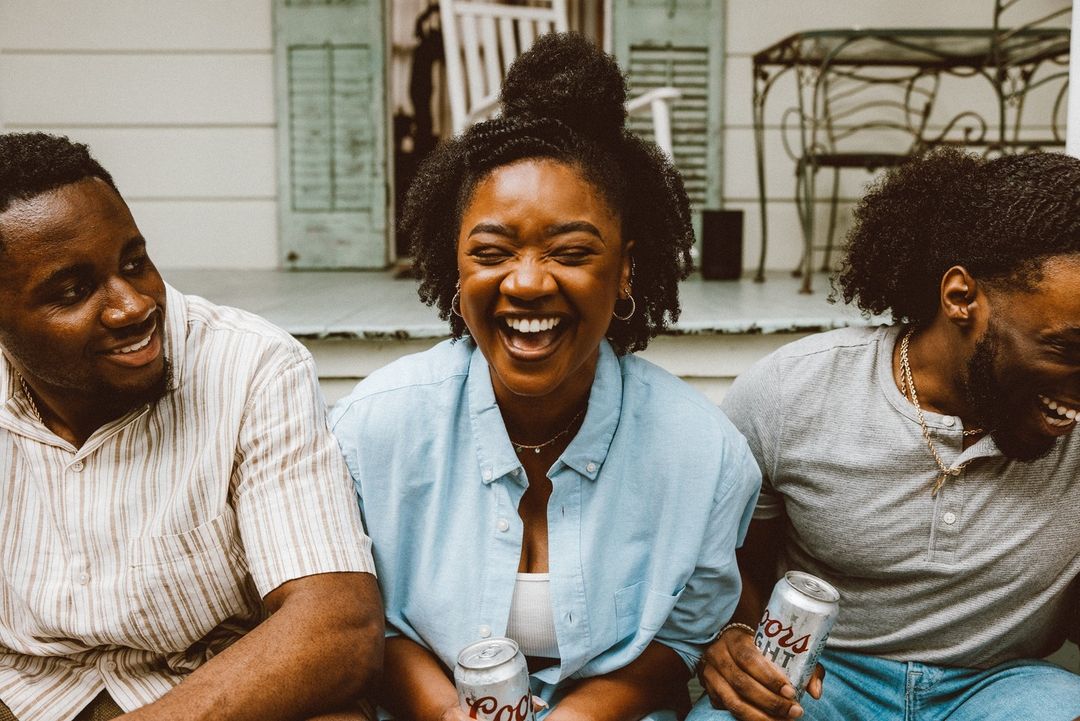 Three young adults sit outdoors, laughing and holding beverages. Two African American men flank an African American woman in the center.