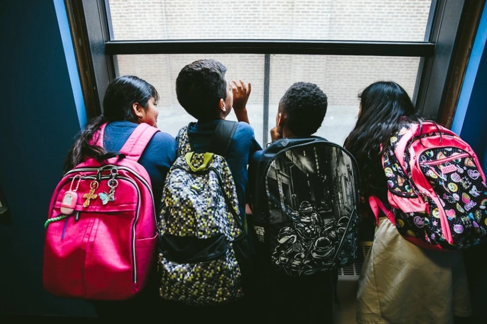 Four children with backpacks stand at a window, rear view. Student group looks out onto a sunny day, waiting to go outside, back to school.