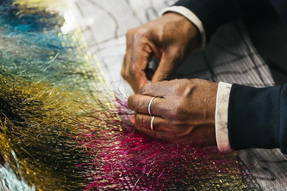 Close-up shot of a person's hands with gold rings, working with colorful, reflective fiber strands on a patterned surface indoors.