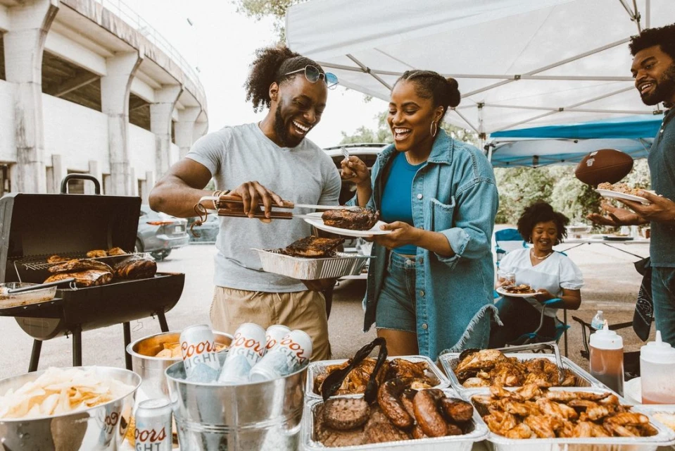Friends grilling food at an outdoor barbecue, laughing together with plates of meat, cans of beer, and food in containers visible.