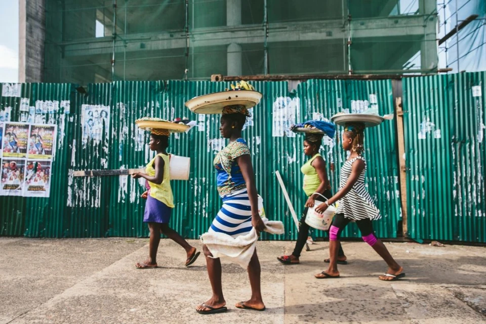 Four African women walk in a line carrying trays on their heads, wearing colorful clothing, on an urban street with a corrugated fence.