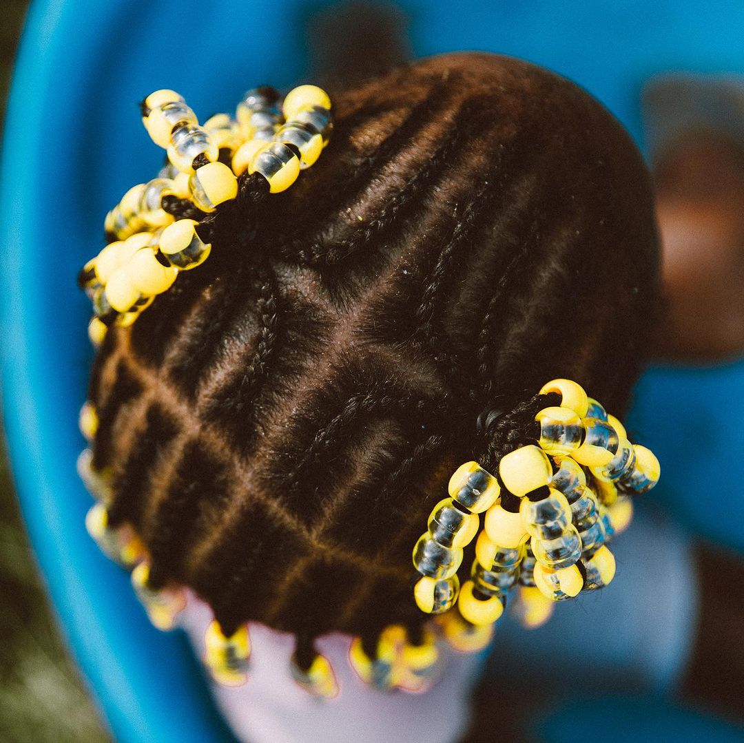 Close up of the braided hair of a child, styled with yellow and black beads at the ends, with a blurred blue and green background.