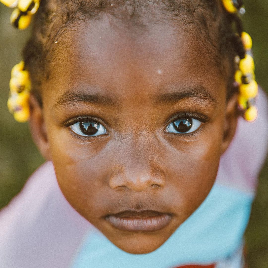 Close-up portrait of a young African girl with beaded braids looking up at the camera in an outdoor setting with soft, natural lighting.