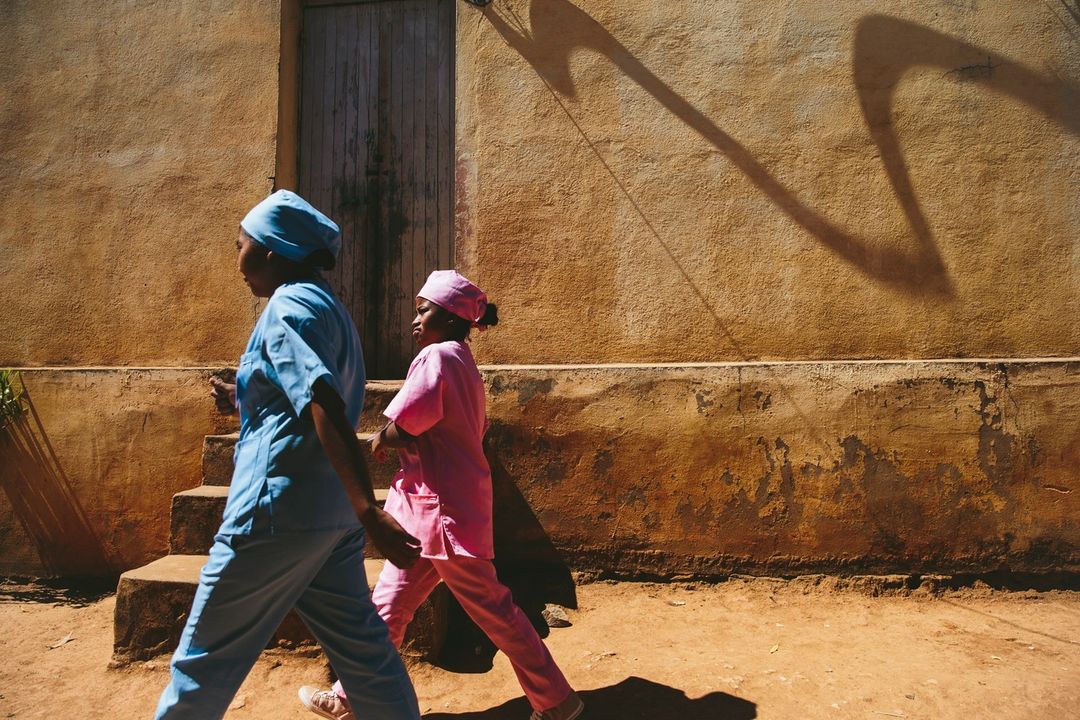 Two healthcare workers, one in blue scrubs and one in pink, walking outdoors alongside a textured, tan-colored wall with shadows.