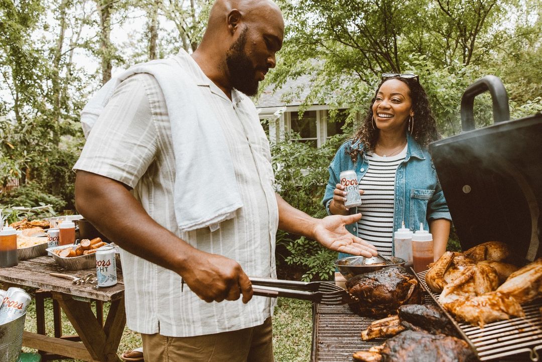 African-American man grilling meat outdoors, while an African-American woman watches and smiles. They are surrounded by a green, leafy backyard.
