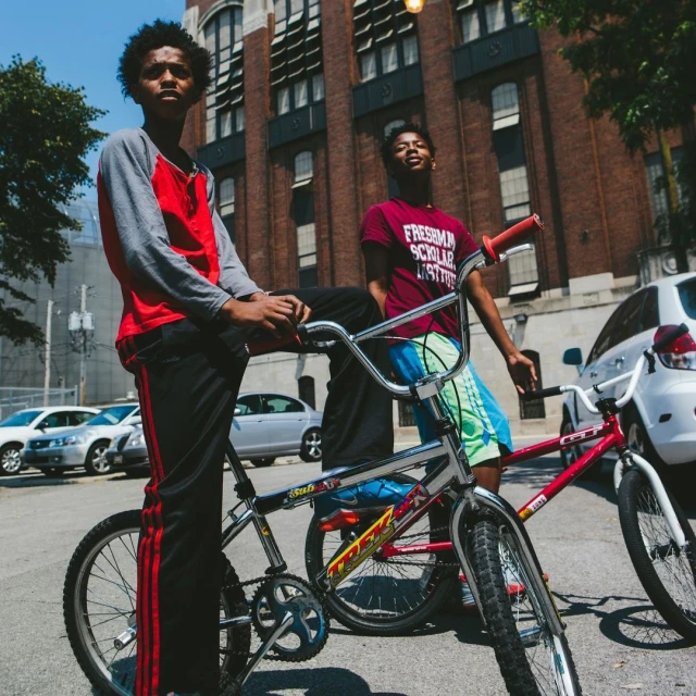Two African-American teenage boys with bikes stand on a street in front of a brick building with parked cars on a sunny day.