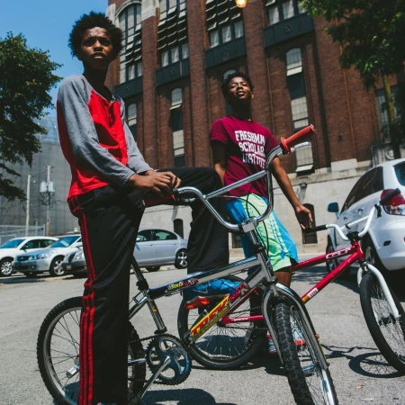 Two African-American teenage boys with bikes stand on a street in front of a brick building with parked cars on a sunny day.
