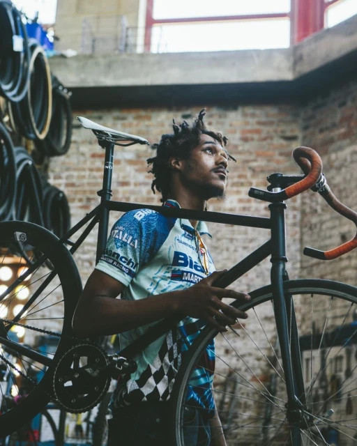 Man carrying a black bicycle in an indoor workshop with bicycle tires and brick wall. The man is wearing a cycling jersey.
