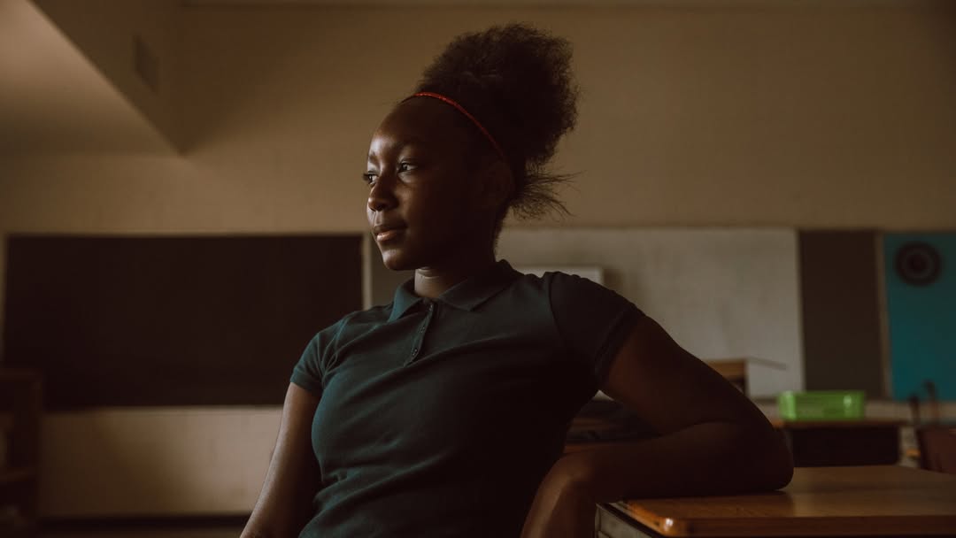 Portrait of an African-American teen girl in a dark-green polo shirt, sitting in a classroom with a chalkboard and desk.