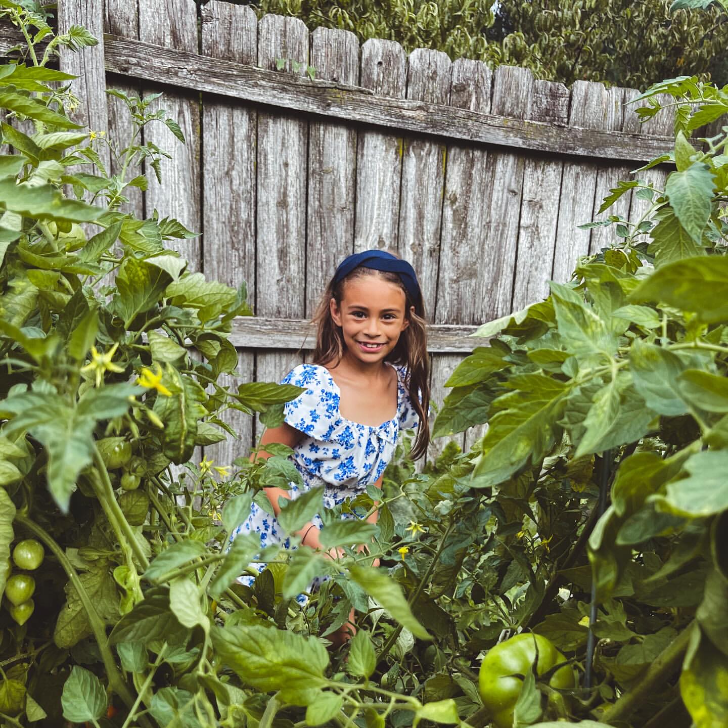Smiling girl in a blue and white dress stands amid lush green tomato plants in a garden with a weathered wood fence backdrop.
