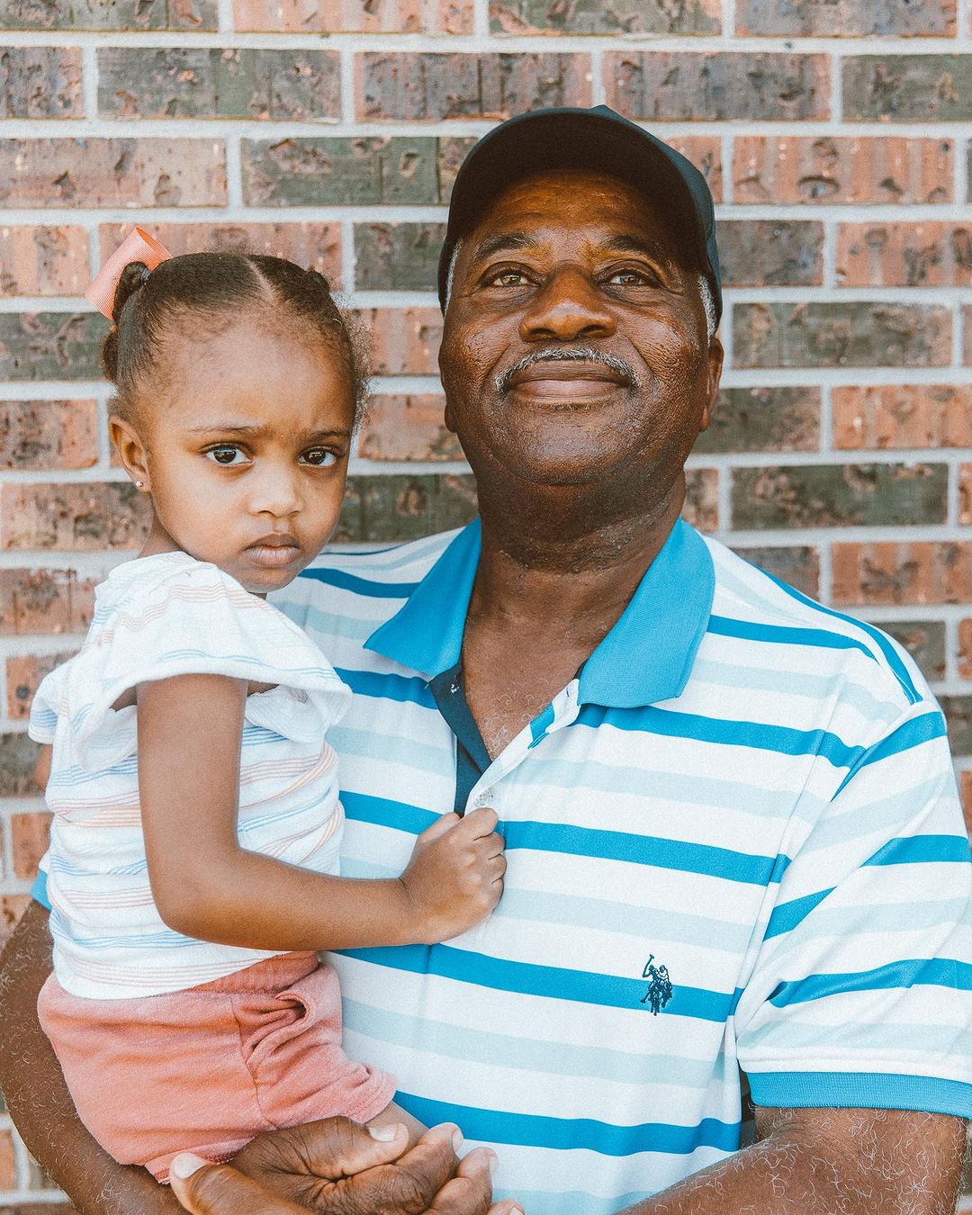 An African-American man holds a young girl in front of a brick wall, showcasing a heartwarming family moment with a close, loving embrace.