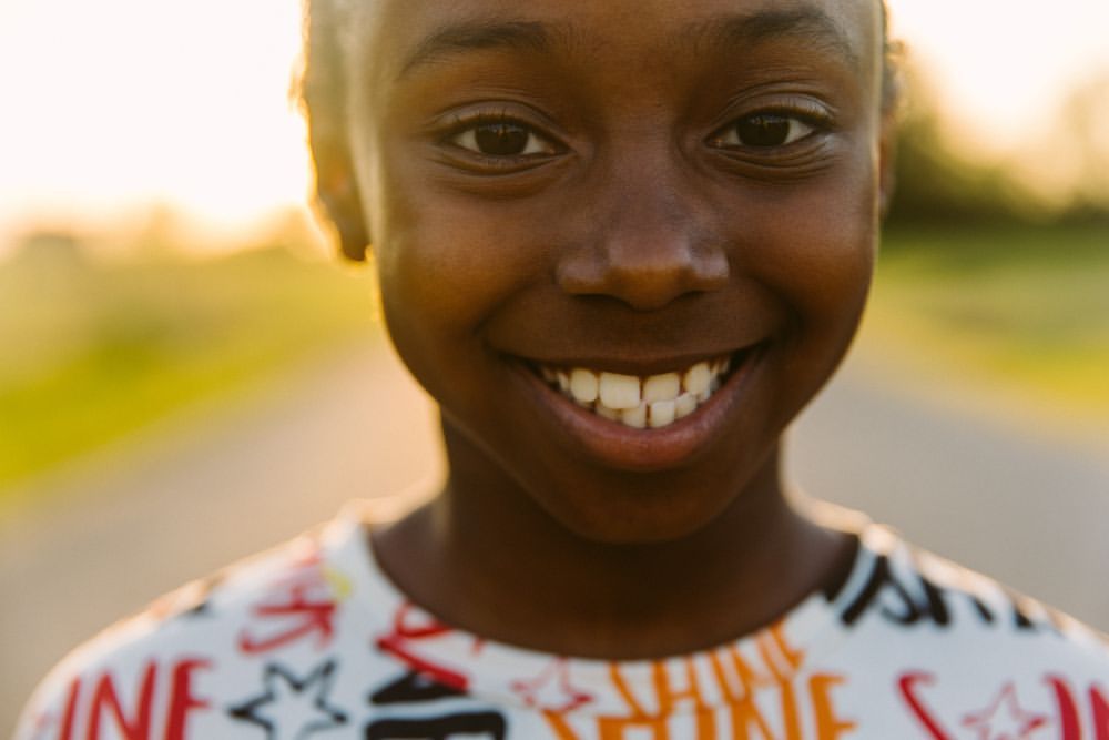 Close-up portrait of a smiling Black child with a blurred background of a path and trees, captured in warm, natural light.