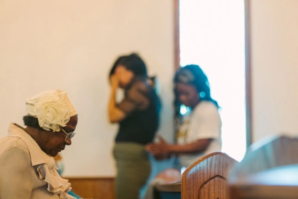 Three women of color gather indoors, with an elderly woman in a white hat and glasses kneeling in the foreground. Natural light.