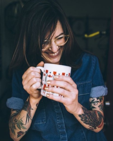 Woman with eyeglasses and arm tattoos smiling while holding a white patterned coffee mug in San Francisco