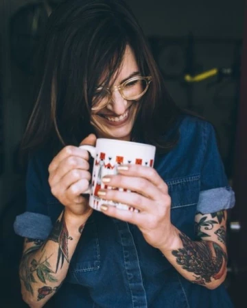 Woman with eyeglasses and arm tattoos smiling while holding a white patterned coffee mug in San Francisco