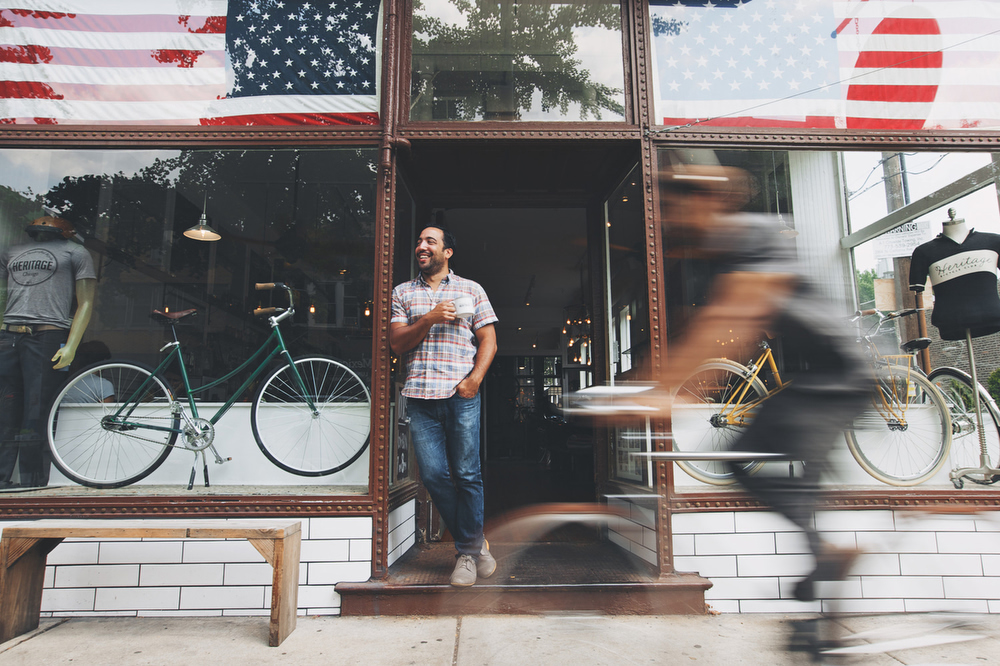 Adult male standing in the doorway of a bicycle shop, with bicycles, bike accessories, and an American flag visible.