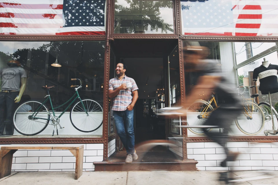 Adult male standing in the doorway of a bicycle shop, with bicycles, bike accessories, and an American flag visible.