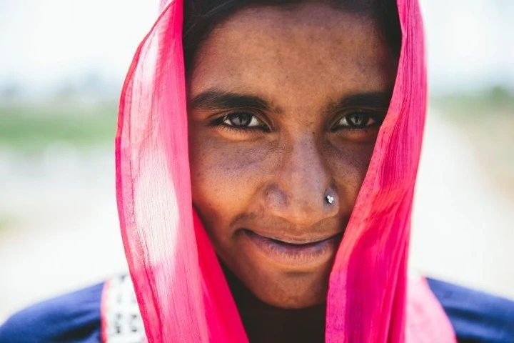 Close-up portrait of a smiling dark-skinned woman wearing a bright pink headscarf, set against a blurred, sunny outdoor background.
