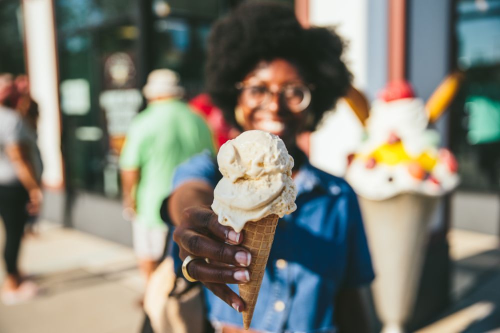 Close-up of a smiling woman holding a melting double scoop ice cream cone outdoors in front of an ice cream store on a sunny day.