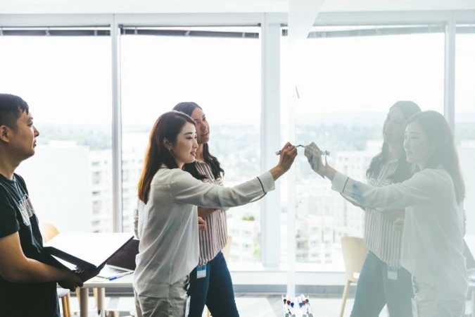 Group of Asian business professionals collaborating in a brightly lit office, writing on a glass wall with city views.