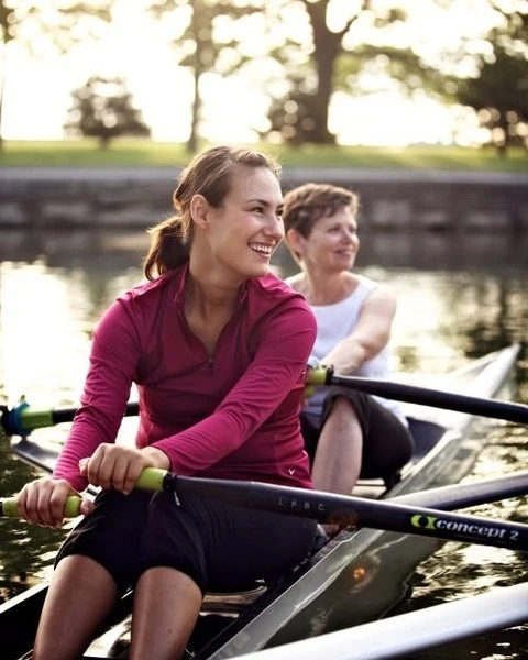 Two women rowing a scull on a river, wearing sporty tops and shorts. Trees and sky in background, with sunlight reflecting off water.
