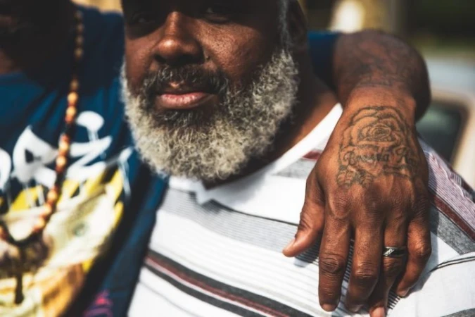 Close-up portrait of a black man with a graying beard, wearing a striped shirt, with an arm draped over his shoulder showing a hand tattoo.