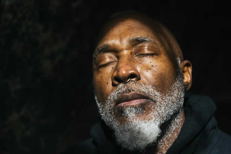 Close-up portrait of an African-American man with closed eyes, gray beard, and nose ring, captured in a dark, moody setting with soft lighting.
