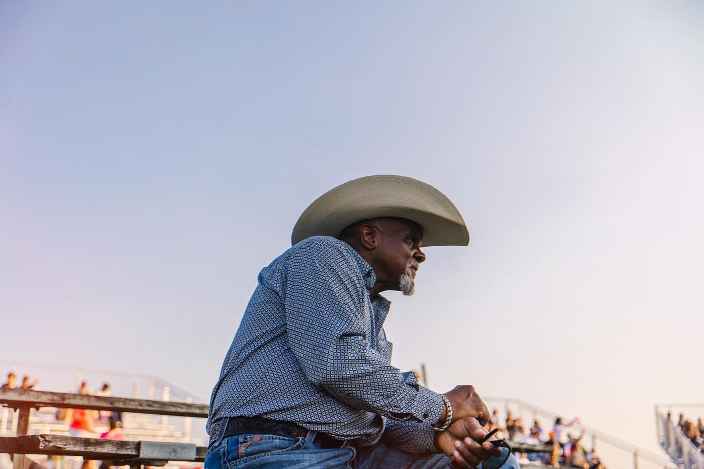 An African-American cowboy sits in the stands wearing a cowboy hat, collared shirt and jeans against a blue sky.