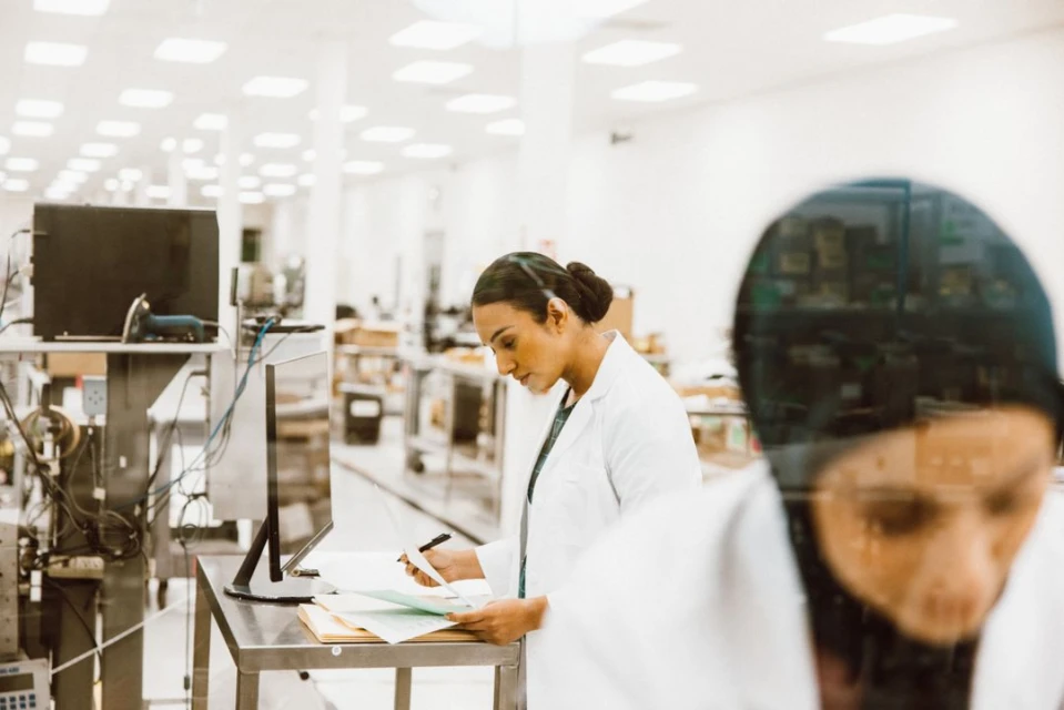 Two healthcare workers in lab coats are recording information in a bright, clean laboratory setting, focused on medical data and research.