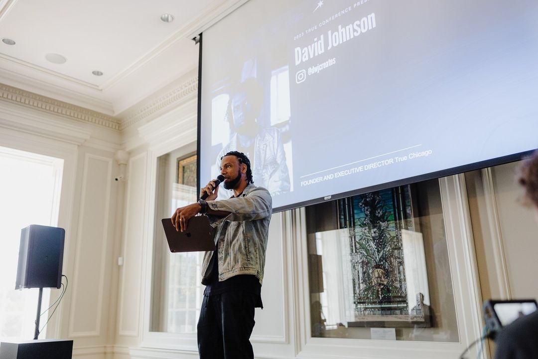 African American man presenting at conference, holding microphone and laptop in indoor hall with a large screen in the background.