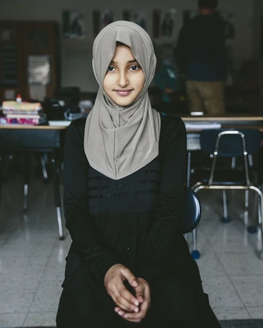 Portrait of a young Muslim woman wearing a hijab and a black dress, seated in a classroom, looking directly at the camera with a smile.