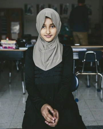 Portrait of a young Muslim woman wearing a hijab and a black dress, seated in a classroom, looking directly at the camera with a smile.
