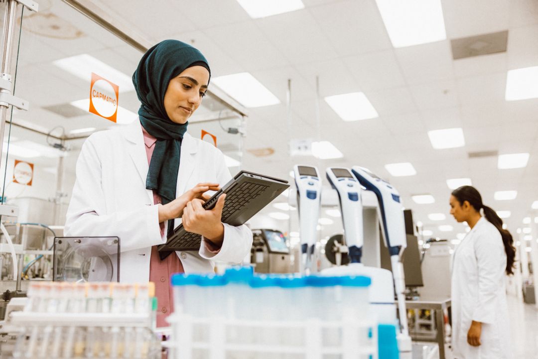 Female scientist in hijab uses a tablet in a bright laboratory setting with test tubes and lab equipment visible in the background.