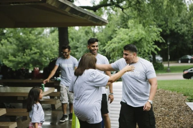 Outdoor gathering featuring a group of people of varying ages, standing and interacting in a park-like setting with trees and picnic tables.