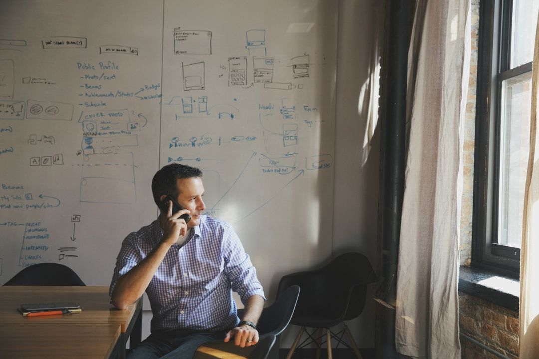Man in blue checkered shirt on phone in office setting, whiteboard with diagrams, natural light from window creates shadows.