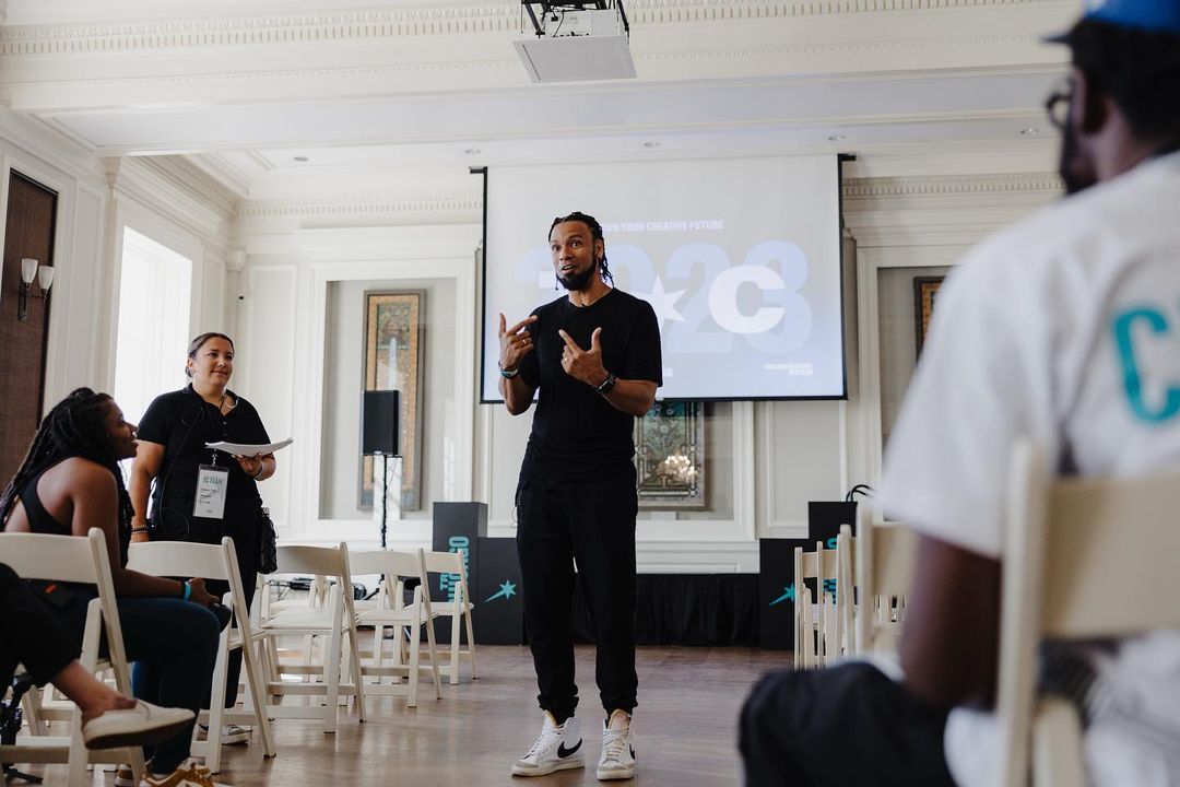 Speaker presents to audience in a bright room with large window, wearing black tshirt and pants, white sneakers, on the stage, indoor.