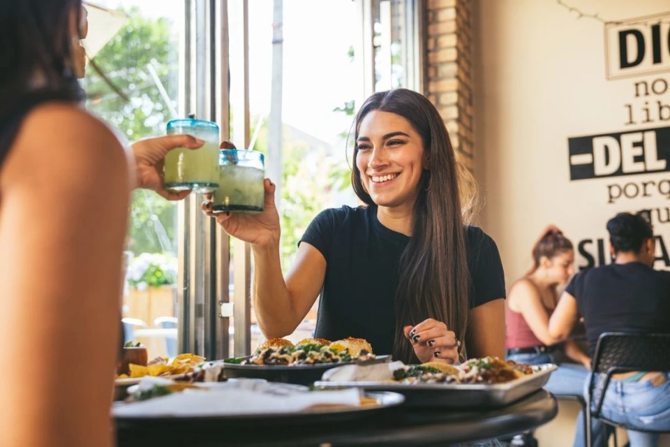 Two women toasting with lime drinks at a restaurant. One woman smiles. Food, plates, and silverware are on the table.