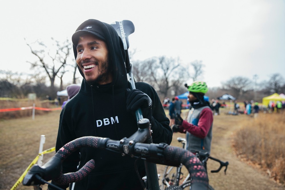 A smiling man in a hooded sweatshirt carries a bicycle on his shoulder at an outdoor cycling event with spectators and tents in the background.