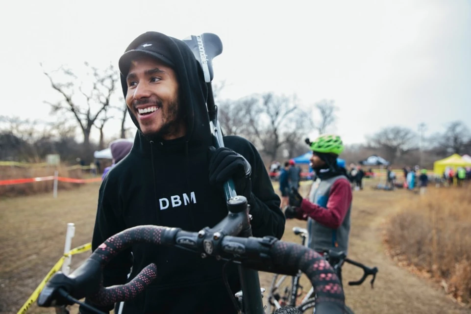 A smiling man in a hooded sweatshirt carries a bicycle on his shoulder at an outdoor cycling event with spectators and tents in the background.