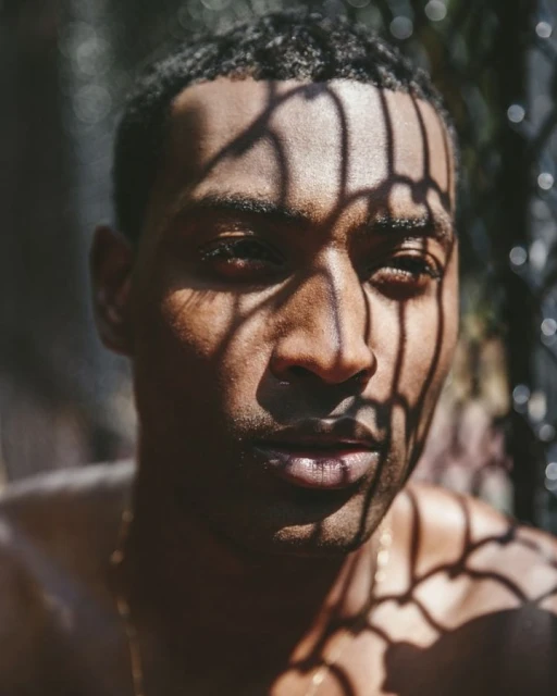 Close-up portrait of a young adult male with dark hair and light brown skin, featuring shadows cast by a geometric structure.