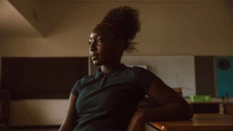 Black teenage girl sitting in a dimly lit classroom wearing a dark polo shirt and headband, looking thoughtfully away.
