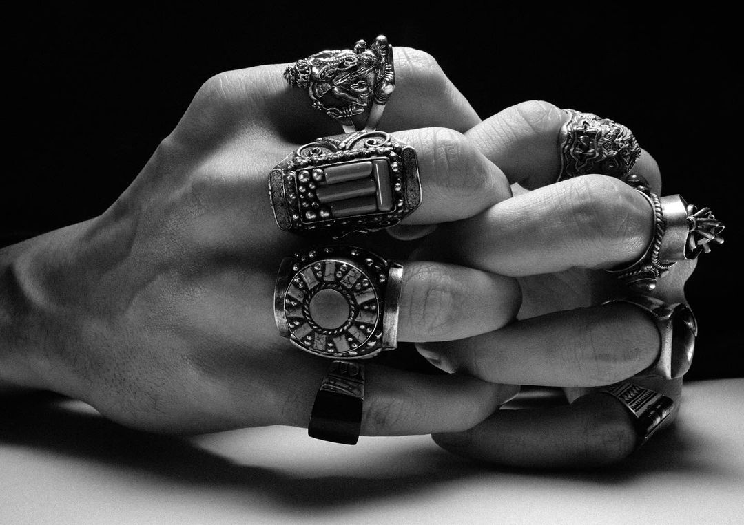 Close-up black and white studio shot of a hand adorned with multiple detailed rings, set against a dark background.