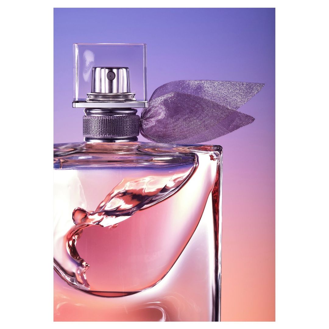 Close-up studio shot of a square perfume bottle with pink liquid, adorned with a translucent ribbon, against a graded purple and orange background.