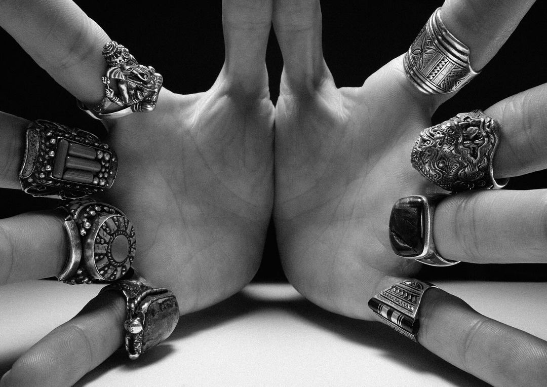 Two hands symmetrically display an array of detailed silver rings on a white surface with a dark background, showcasing intricate jewelry.